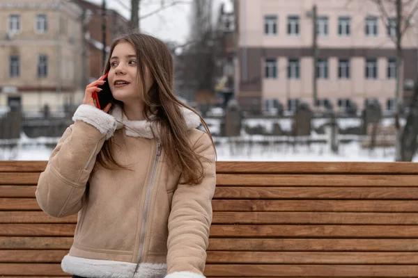 Young woman in city talks on phone and sits on park bench. Girl in beige sheepskin coat rests outside in winter.