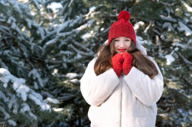 Kırmızı şapkalı ve karlı arka planda eldivenleri olan gülümseyen genç bir kadının portresi. Mutlu kız kış ormanı.