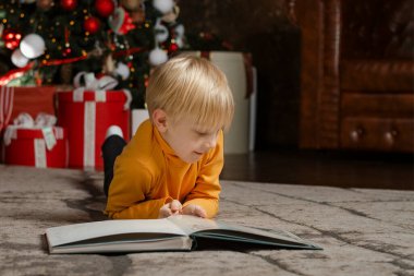 Sarışın çocuk yılbaşı ağacı arka planında kitapla yerde yatıyor. Beş ya da altı yaşındaki çocuk kitap okur.