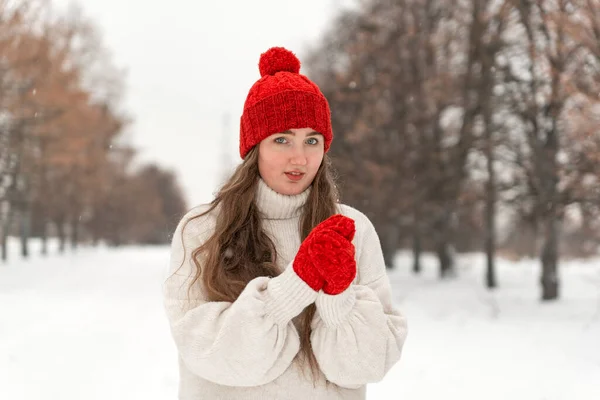 冬の公園の路地に赤いニット帽とミトンとウールのセーターの美しい若い女性の肖像 — ストック写真
