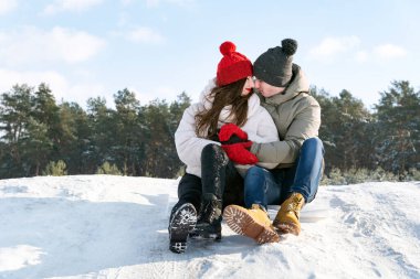 Genç aşıklar, kış ormanı ve mavi gökyüzünün arka planında oturup kucaklaşırlar. Kış güneşli gün.