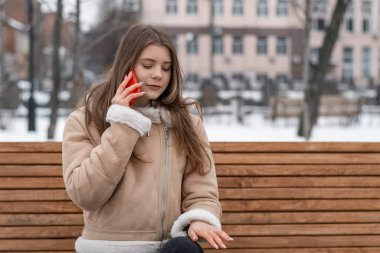 Genç bir kadın kış parkında telefonla iletişim kuruyor. Koyun derisi ceketli bir kız parkta bankta oturan bir alet kullanıyor..