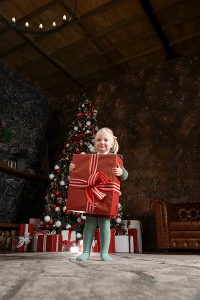 Little girl with big gift on xmas tree background. Cute blonde child girl with huge red gifts box
