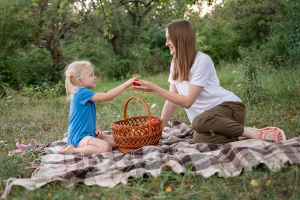Pretty little girl gives apple her mother or sister, on clearing in forest. Summer family picnic of mom and girl in park.