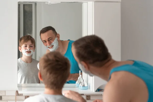 Dad and son in shaving foam on faces have fun in bathroom. Morning facial hygiene.