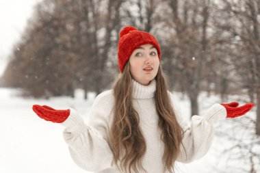 Long-haired young woman in red hat and mittens outdoor. Portrait of happy girl walking in winter park