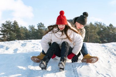 Young guy and a girl slide down the snow slide against the background of the forest and the blue sky