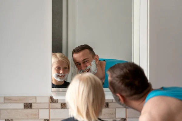 Young dad and little boy with shaving foam on face look in bathroom mirror. Dad and son shave and having fun.