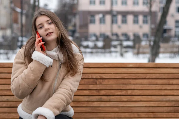 Young pretty woman communicates on phone sitting on bench in winter park . Girl in stylish sheepskin coat uses gadget outdoor.