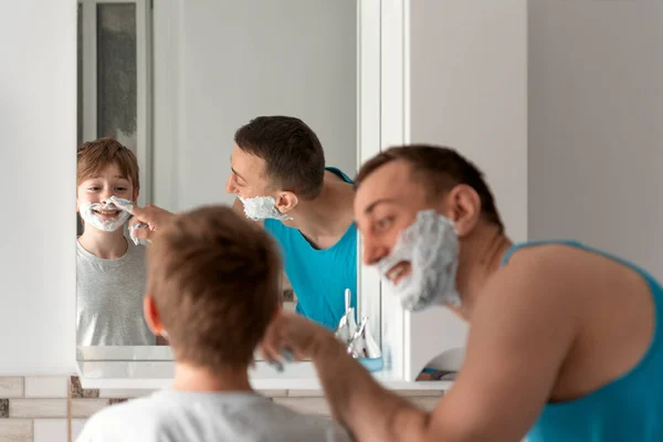 Young dad and his son in shaving foam on faces have fun in bathroom. Morning facial hygiene, time together.