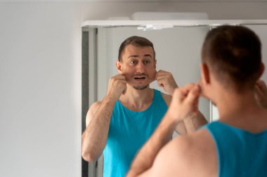 Young man grimaces and makes faces to his reflection in mirror in bathroom. View over the shoulder