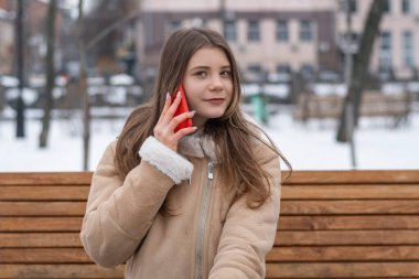 Beautiful brunette girl in sheepskin coat sits on bench in winter park and talks on smartphone. Serious young woman with gadget