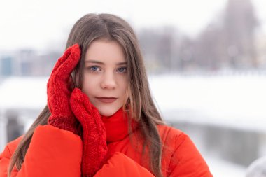 Close up portrait of blue-eyed eyes girl in red warm jacket and mittens in winter outside. Youth warm clothing.