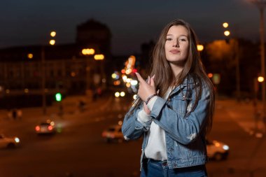 Happy girl student walks around the evening city. Young brunette woman in denim clothes on evening city background