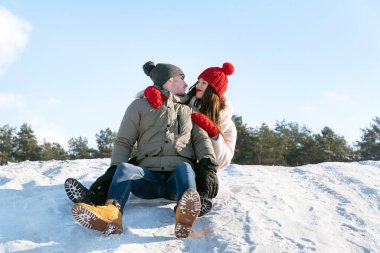 Young guy and a girl sit on the snow against the background of the forest and the blue sky. Couple outdoors.