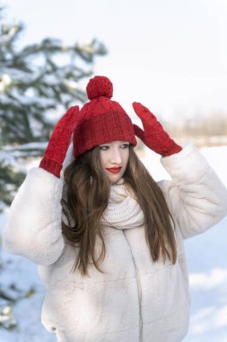 Genç bir kadın kış parkında şapkasını karlı arka planda düzeltiyor. Dikey çerçeve. Güneşli kış günü.