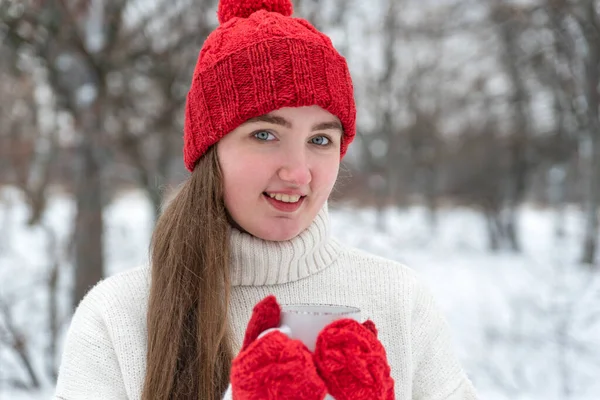 Portrait of smiling woman in red wool hat and mittens holding cup of hot drink in outside.