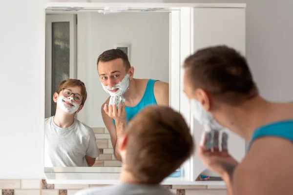 Father and son shaving in bathroom together and having fun. Dad and son in bathroom in front of mirror.