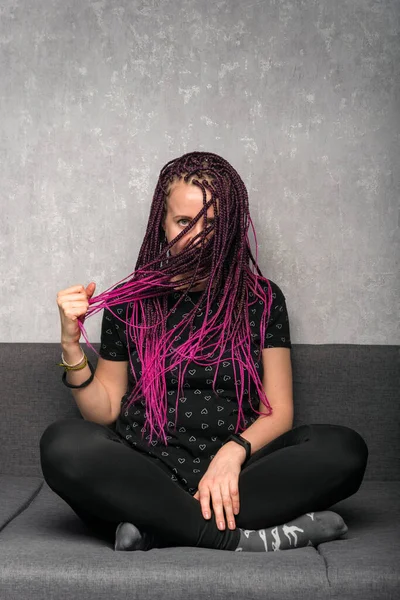 Young caucasian woman with color dreadlocks sits on couch and touches her hair. Vertical frame