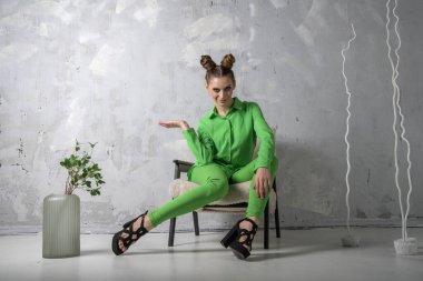 Smiling girl with interesting hairstyle and makeup poses in studio on gray background. Young woman in green suit on chair. Place for your text.