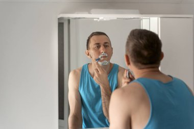 Young man with shaving foam on face removes his beard with razor. Reflection in mirror. Face care.