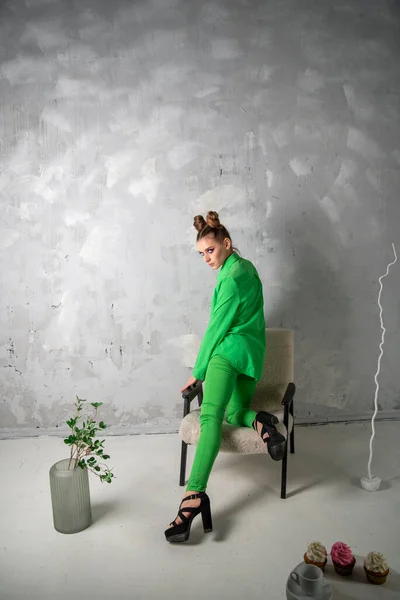 Beautiful girl in green shirt and trousers on chair on gray wall background. Stylish woman photographed in studio. Vertical frame.