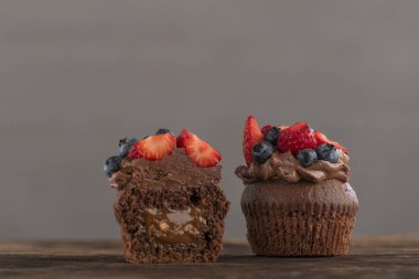 Two fresh cupcakes with chocolate and berries. Cut muffin with cream and fruits on the wooden table. Gray background.
