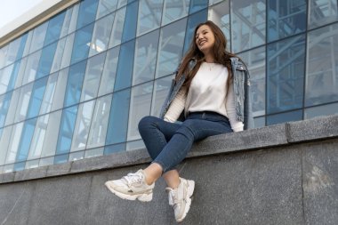 Yaz kenti caddesinde gülen şık bir kız. Güzel öğrenci kız modern binanın yanında oturuyor..