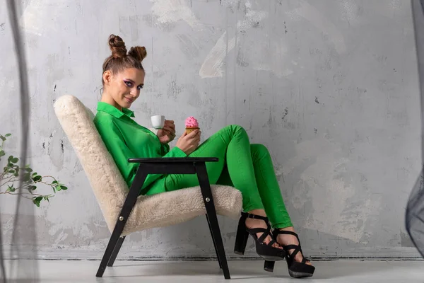 stock image Extraordinary girl in trouser green suit with cup of coffee and muffin sits on chair in studio. Model with interesting hairstyle and bright makeup poses on gray background.