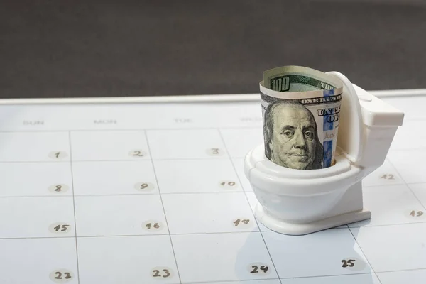 Concept of throwing money down the toilet. Small toy white toilet closet with cash dollars on calendar background.