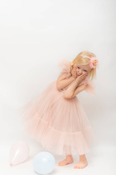 Cute little girl in lush chiffon dress funny poses. Little girl with blonde hair isolated on white background. Vertical frame.