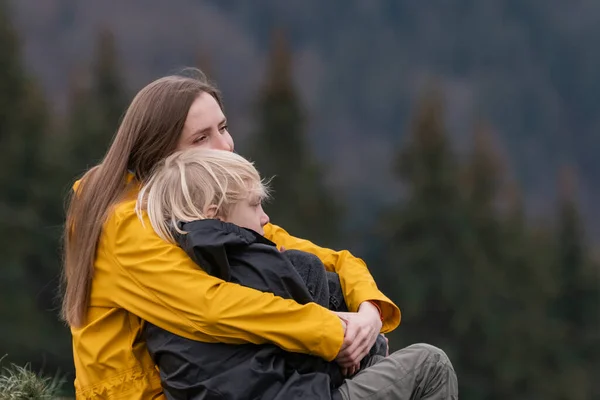 Portrait of young mom and son outside. Mother hugs her child son sitting on halt in woods. Happy family. Hike in the fall