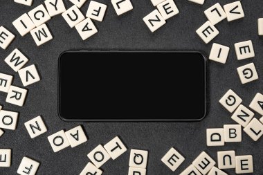 Gri arkaplanda İngiliz alfabesinin beyaz harflerinden yapılmış çerçevesi boş ekranlı siyah akıllı telefon. Boşluğu kopyala