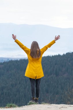 Genç bir kadın ellerini kaldırmış, dağın yamacında dikiliyor. Sarı ceketli kadın turist ya da dağlarda yağmurluk.