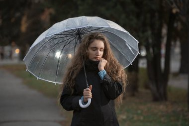 Uzun saçlı kara kara düşünen kız yağmurlu havada şeffaf şemsiyeyle sonbahar parkında yürüyor.