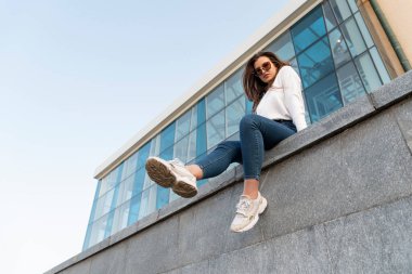 Genç esmer kız korkulukta bacaklarını sallayarak oturuyor. Şehir. Çekici stil sahibi kadın şehir sokaklarında dinleniyor.