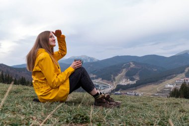 Sarı yağmurluklu mutlu yürüyüşçü portresi. Bir fincan sıcak çayla birlikte. Dağlara bakan bir kadın.