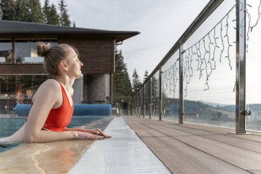 Bun saç stiline sahip kırmızı mayo giyen genç bir kadın açık havuzda güneşli bir günün tadını çıkarıyor. Yan görünüm