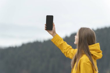 Dağlarda sarı ceketli genç bir kadın elinde boş siyah bir ekranla telefonu tutuyor. Uzayı kopyala Model