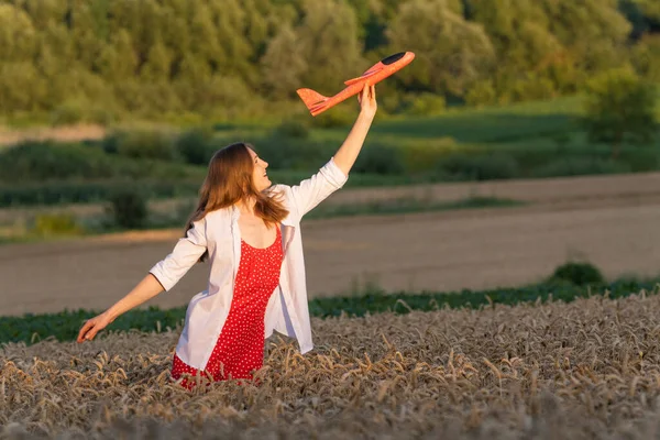 Beautiful young woman with toy airplane in hands in middle of summer wheat field, in red dress and white shirt Concept of cheap air travel.