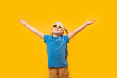 Sarı arka planda güneş gözlüklü ve mavi tişörtlü sarışın kız portresi. Elleri havada küçük bir kız.