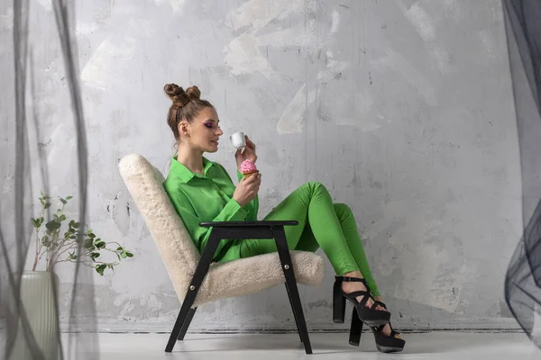 Extraordinary girl in trouser green suit with cup of coffee and muffin. Model with hairstyle and bright makeup poses on gray background.