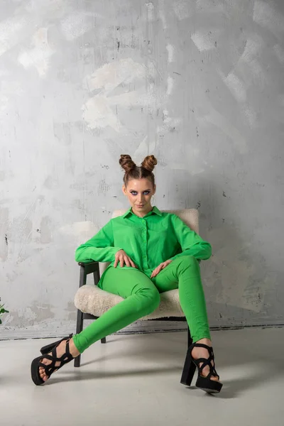 stock image Slender girl with evening makeup and hairstyle poses on chair in studio. Lady in green pantsuit and heels on gray background.