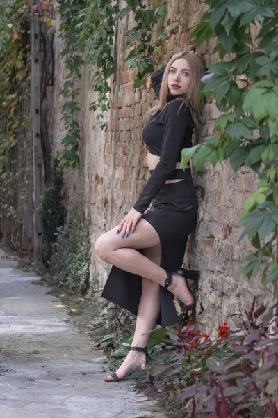 Girl model in black outfit near old wall outdoors. Young beauty woman poses for camera near brick wall. Vertical frame.