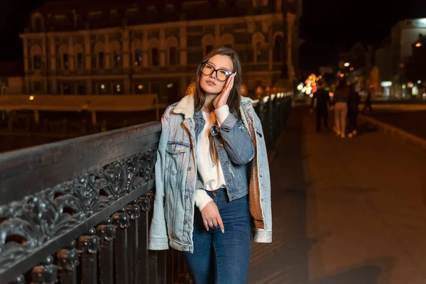 夜の街の背景に眼鏡やデニムスタイルの魅力的な若い女性 スタイリッシュな女の子が夜の街を歩く — ストック写真