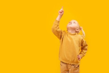 Sarı arka planda sarı takım elbiseli kızın stüdyo portresi. Çocuk elini kaldırdı ve parmağını kaldırdı. Metin için boşluk