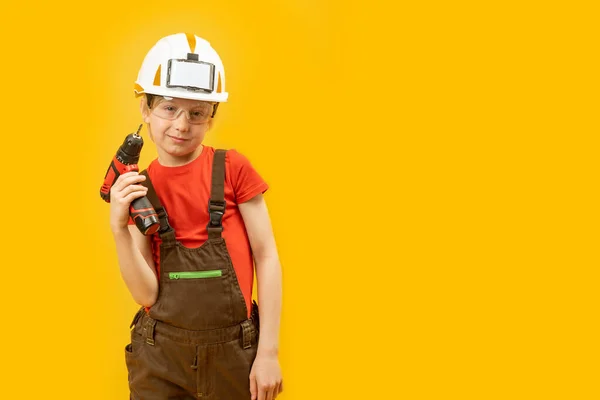 建築家としての子供 白いヘルメットの少年は 保護メガネは作業ジャンプスーツを着用し ドリルを保持します 黄色の背景に隔離された — ストック写真