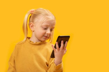 Şaşkın küçük kız cep telefonundan izliyor. Telefonu olan çocuk. Sarı arka plan. Akıllı telefonlu bir çocuğun portresi