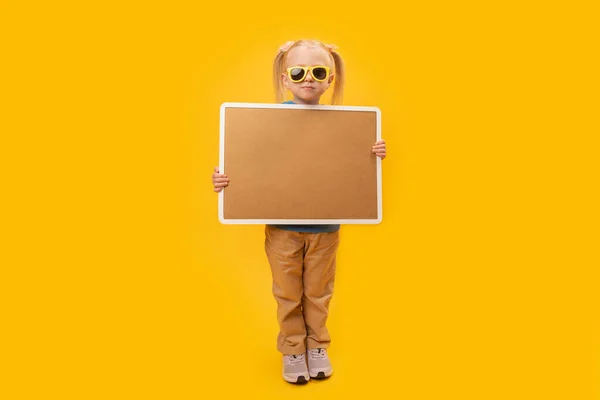 stock image Full-length studio portrait of girl in sunglasses holding an empty cork board. Space for your text. Isolated on yellow background