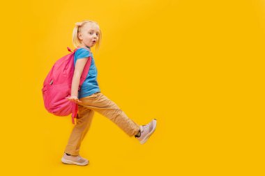 Büyük okul çantalı küçük kız bacağını kaldırıyor ve sarı arka planda koşuyor. İlkokul öğrencisi okula hazır. Boşluğu kopyala.